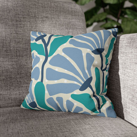 A square throw pillow with a bold botanical pattern in teal, light blue, and cream, resting on a textured gray fabric armchair.