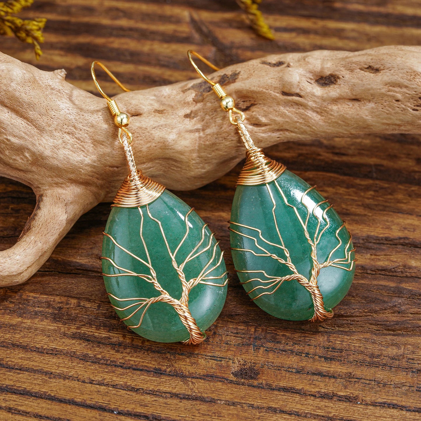 A pair of tree-of-life drop earrings with green teardrop-shaped stone pendants, displayed on a piece of driftwood over a wooden surface.