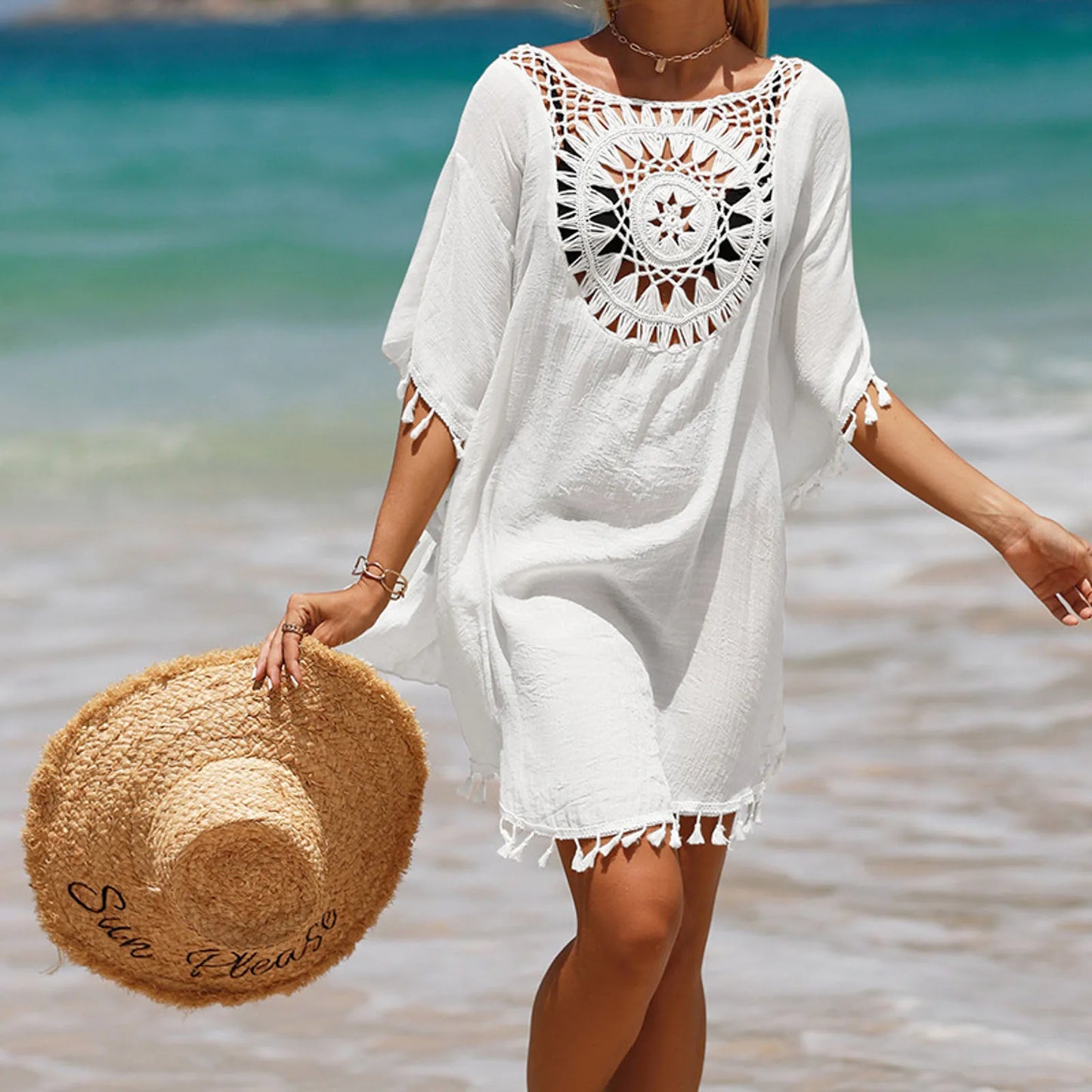 Woman in a white beach cover-up with crochet details holding a sun hat on a beach.