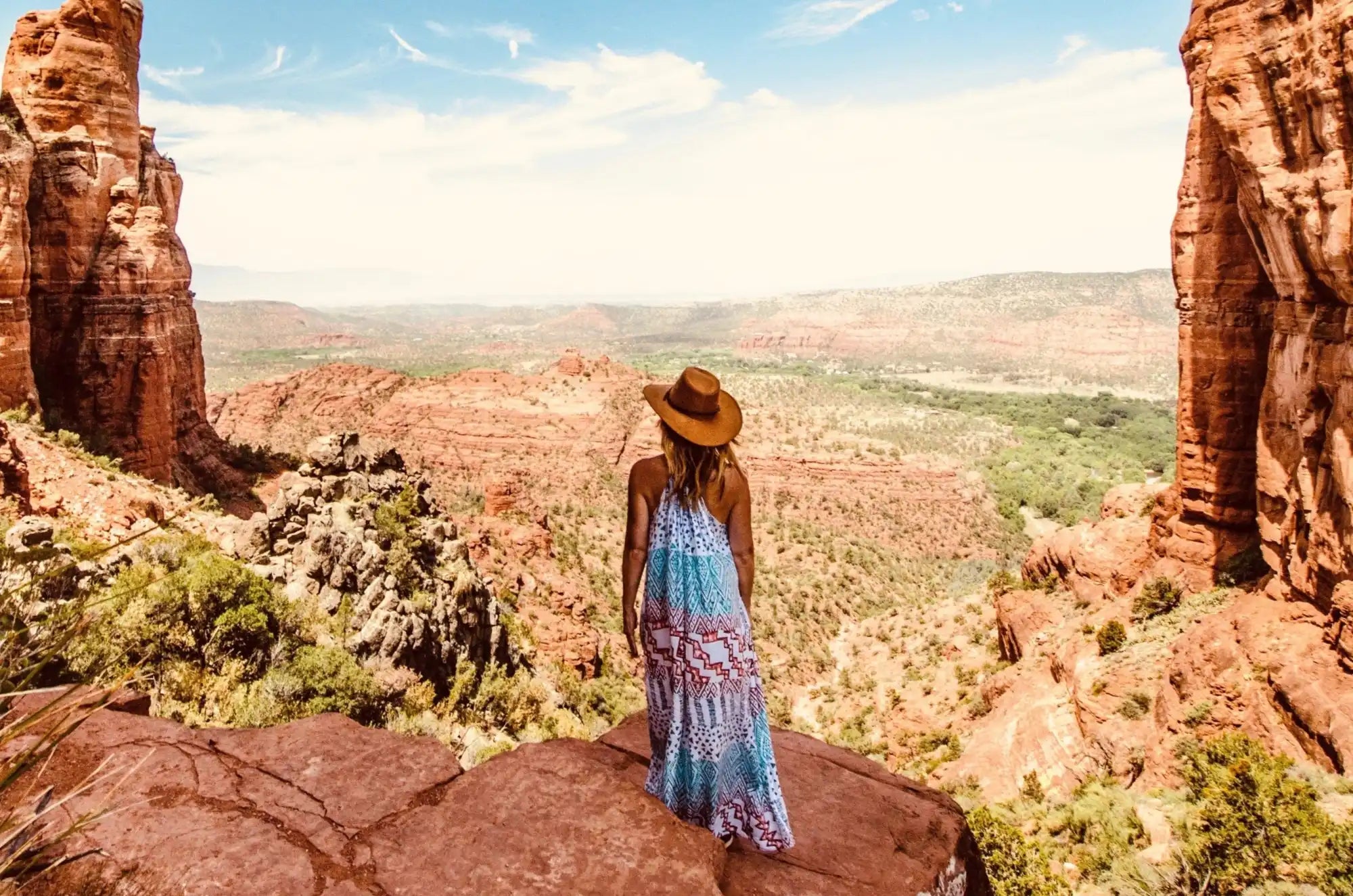 Woman is standing on a ledge looking out over a desert valley. She is wearing a sundress and a straw hat.